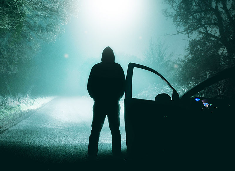 A lone, hooded figure standing next to a car looking at an empty