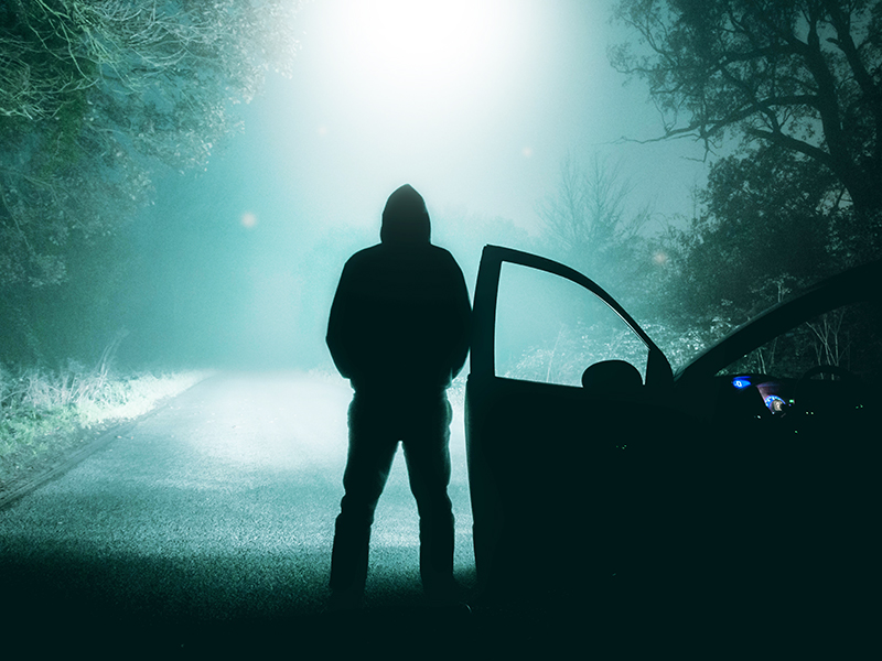 A lone, hooded figure standing next to a car looking at an empty