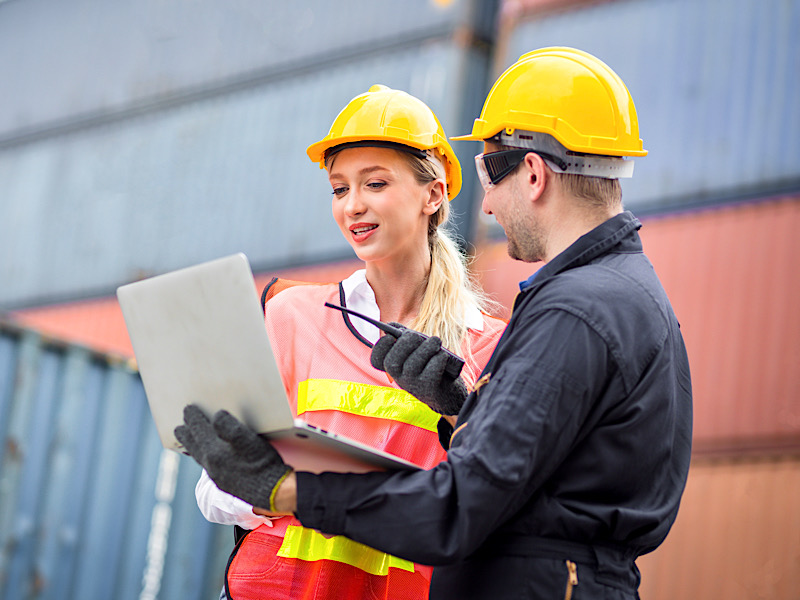 Female dock worker wearing a white helmet yellow standing in a industrial shipping yard she discussing colleagues and control container transportation and export with commercial docks.